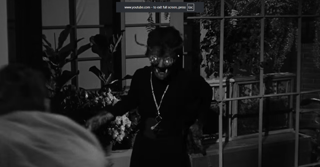 A character in a dark outfit with a grotesque face and glasses, stands in a dimly lit room surrounded by plants.