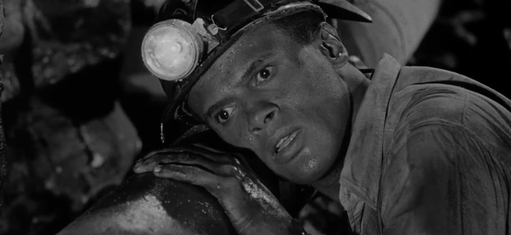 A miner in a helmet with a light looks concerned while leaning against a pipe in a dark underground setting.