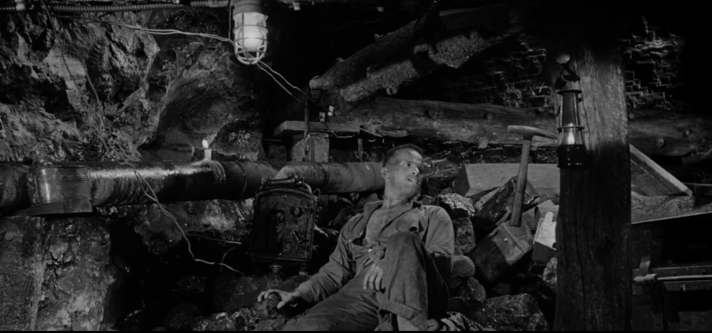 A miner resting on a pile of rocks in a dimly lit underground mine, with a lantern illuminating the scene and mining equipment in the background.