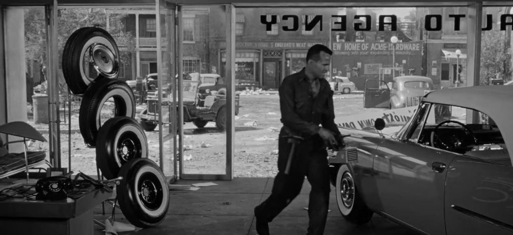 A man in a dark shirt walks through an auto agency, with vintage cars visible outside and stacks of tires in the foreground.