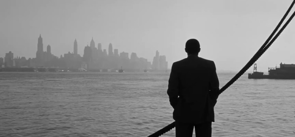 A silhouette of a man in a suit standing by the water, looking toward a foggy city skyline in black and white.