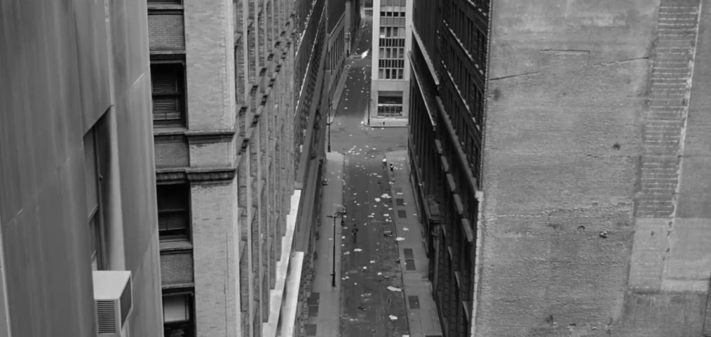 A black and white aerial view of a narrow street between tall buildings, with scattered debris on the pavement.