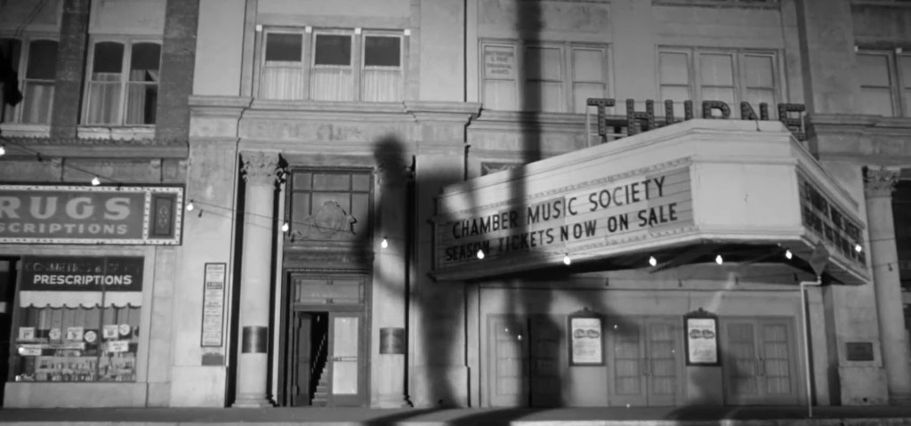 A black and white photograph of a vintage theater façade with a marquee advertising the 'Chamber Music Society' and 'Tickets Now On Sale,' alongside surrounding shops displaying various signs.