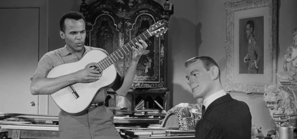 A musician playing a guitar while singing to a ventriloquist dummy in a vintage setting, surrounded by ornate furnishings and artwork.
