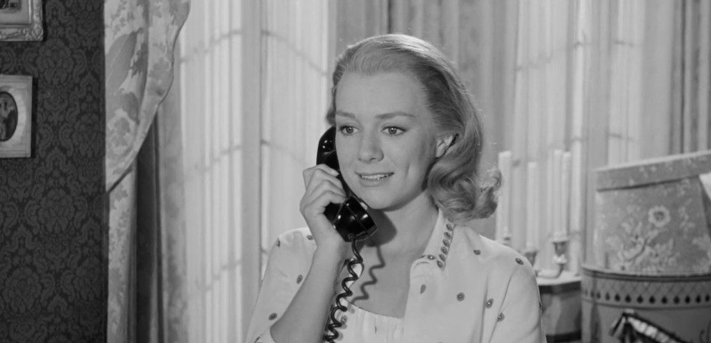 A woman smiling while talking on a vintage black telephone in a softly lit room with curtains and decorative elements.