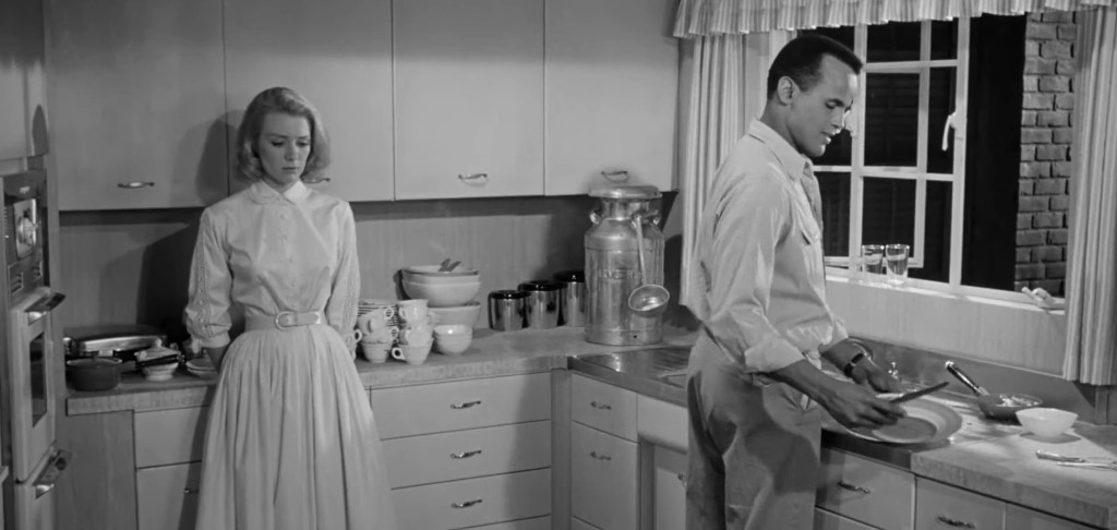 A woman stands silently in a kitchen, looking concerned, while a man washes dishes at the sink.