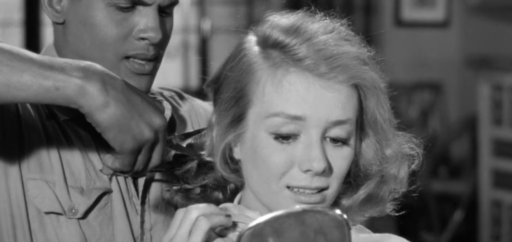 A man is cutting a woman's hair with scissors while she looks at her reflection in a hand mirror, displaying a mix of concern and curiosity.