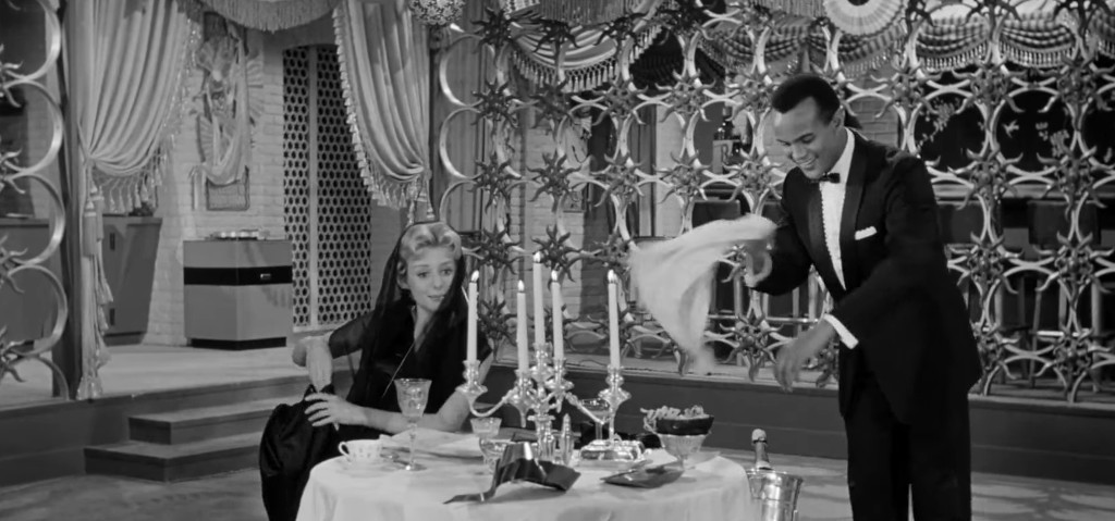 A black and white scene featuring a dining table set with candles and elegant dishes, with a woman sitting and a man in formal attire leaning over the table, appearing to serve or present food.