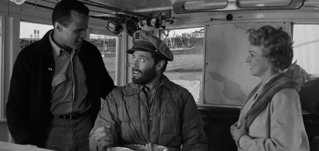 A black and white scene showing three people interacting on a boat; a man in a jacket is talking to a bearded man in a captain's hat, while a woman in a light coat smiles at them.