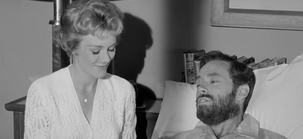A woman in a white sweater smiles gently at a man lying in bed, who has a beard and is wearing a shirt. The scene is set in a simple room with a lamp and books in the background.