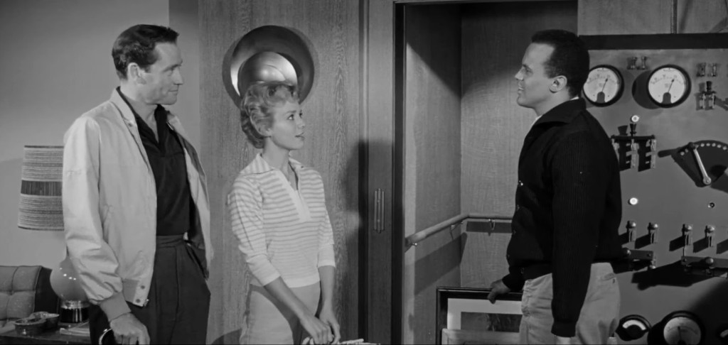 A black and white scene featuring two men and a woman engaged in conversation in an indoor setting, with a modern design backdrop and various equipment visible in the background.