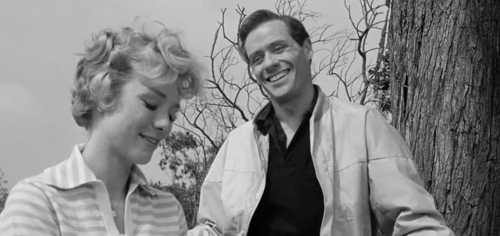 A smiling man in a jacket stands beside a woman with curly hair, who is looking down with a slight smile, against a backdrop of bare trees.