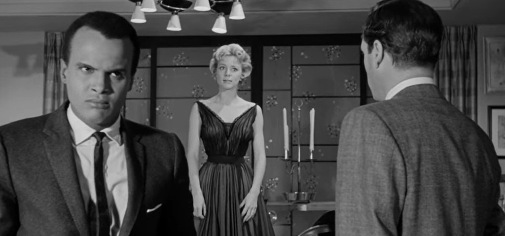 A tense scene featuring a man in a suit with an angry expression, a woman in a black dress standing calmly, and another man in a gray suit looking toward them.