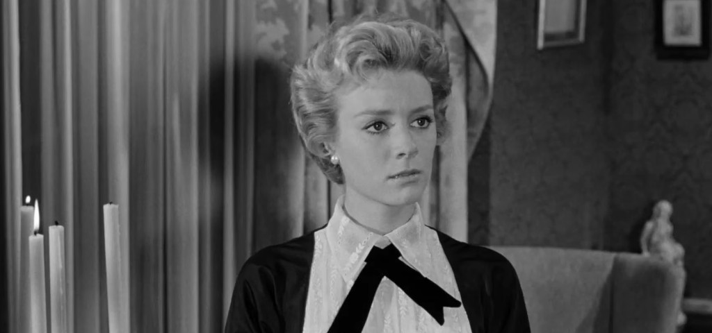 A woman with short, styled blonde hair wearing a black dress with a white collar and bow, looking serious in a black and white setting.