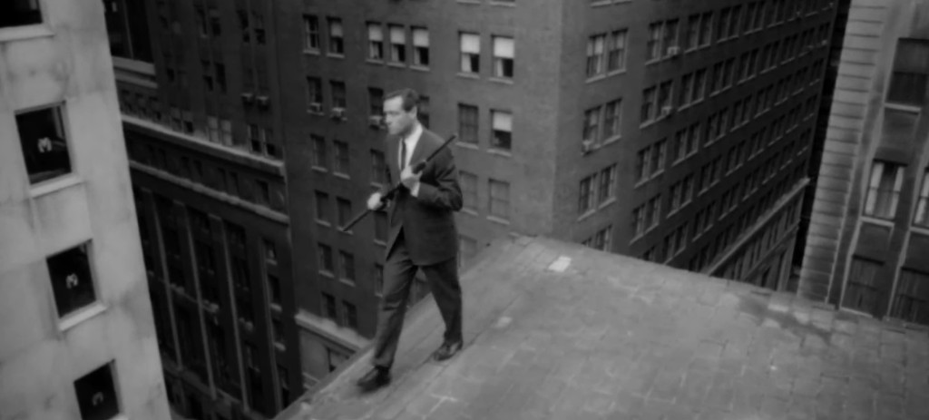 A man in a suit walking carefully along the edge of a rooftop in a cityscape, with tall buildings in the background.