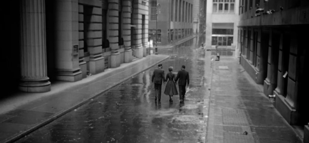 Three figures walking hand-in-hand down a rainy, deserted city street, with wet pavement and tall buildings lining the sides.