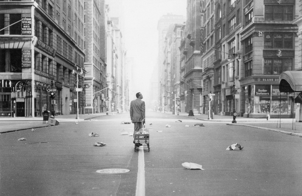 A lone figure stands in the middle of a deserted street, pushing a cart, surrounded by scattered debris and buildings on either side, captured in black and white.