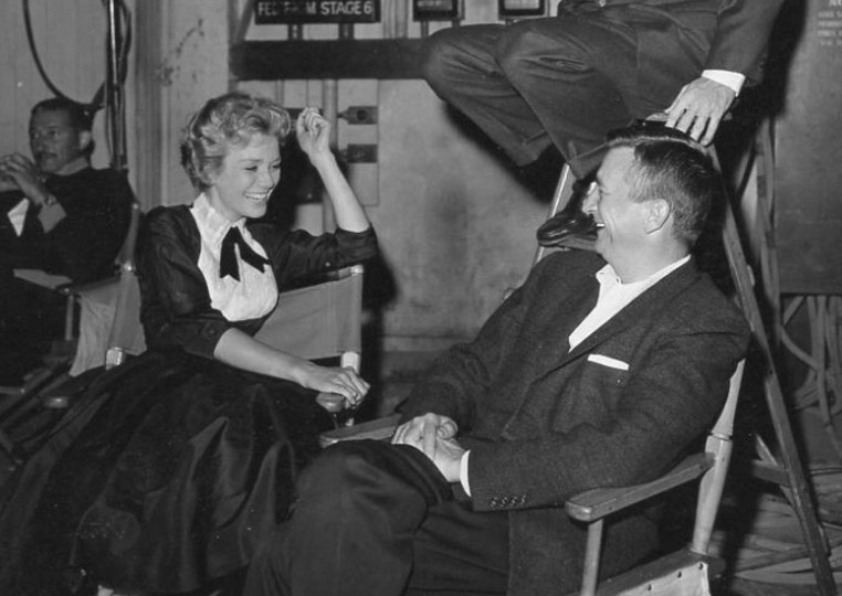 Black and white photo of a woman in a vintage dress laughing while talking to a man in a suit, both sitting in director's chairs on a film set.