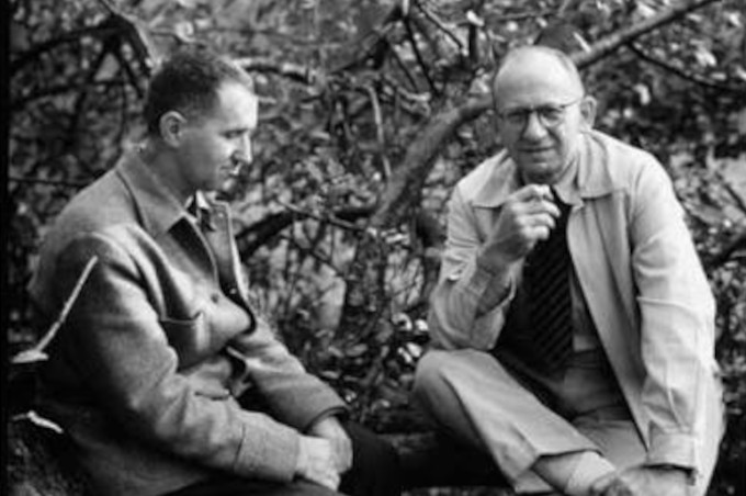 Two men seated on the ground next to a tree, engaged in conversation, in a black and white photograph.
